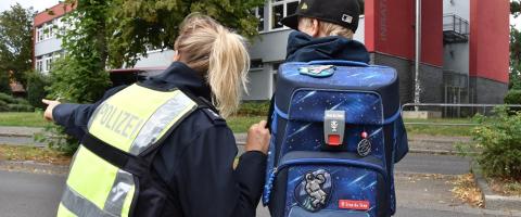 Ein Schulkind steht neben einer Polizistin. Die Polizistin zeigt nach links vorne