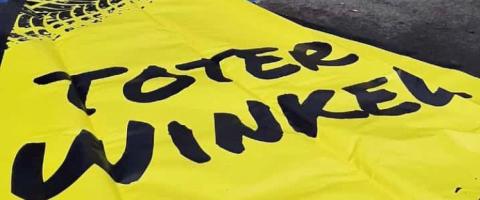 Eine gelbe Plane liegt auf dem Boden. Die Aufschrift "Toter Winkel" ist zu lesen, im oberen Bildrand sind Reifenabdrücke zu sehen