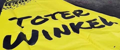 Auf der Straße liegt eine gelbe Plane mit der Aufschrift Toter Winkel. Die Plane hat die Form eines Dreiecks. Im Hintergrund steht ein Transporter