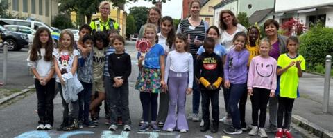 Schulkinder, Lehrer und eine Polizistin stehen auf einer Straße. Im Vordergrund ist der Schriftzug "Achtung Kinder" zu lesen