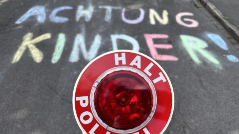 Auf einer Straße ist mit bunter Kreide der Schriftzug "Achtung Kinder!" gemalt. Im Vordergrund des Bildes ist eine Polizeikelle mit dem Schriftzung "Halt Polizei" zu sehen