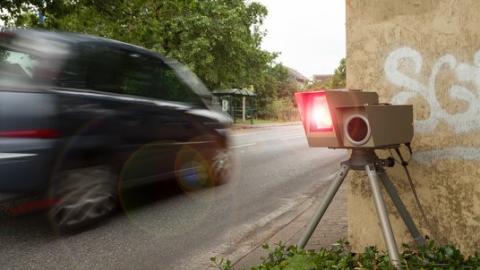 Auf der rechten Bildseite ist eine Radarkamera mit aufblitzendem Rotlicht erkennbar. Links fährt ein Auto durch das Bild