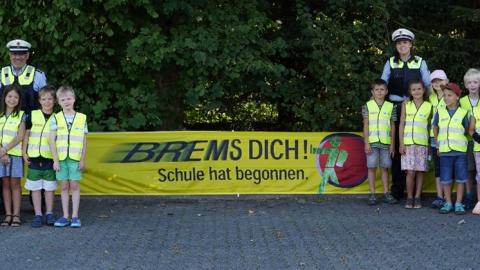 In der Bildmitte hängt ein Plakat "Brems dich! Schule hat begonnen" der Landesverkehrswacht in gelb. Links und rechts stehen Grundschulkinder mit Polizeibeamtinnen und Polizeibeamten
