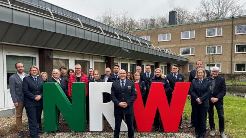 In der Mitte des Bildes stehen die drei Buchstaben NRW in den Landesfarben grün, weiß und rot. Die Mitglieder der Projektgruppe stehen neben und hinter dem Schriftzug, der Projektleiter davor