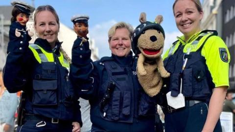 Drei Polizeibeamtinnen stehen in Uniform zur Kamera. Sie tragen auf ihren Armen jeweils Puppen die zur Verkehrserziehung genutzt werden.