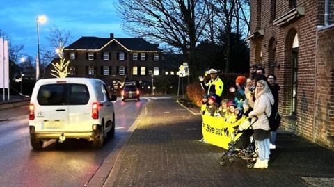 Auf einem Gehweg stehen in der Dämmerung Schülerinnen und Schüler sowie Eltern, alle tragen gelbe Warnwesten und Reflektoren. Sie halten ein Banner, den Schriftzug kann man nicht erkennen. Im linken Bildrand fährt ein Auto vorbei