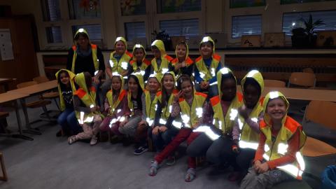 Eine Gruppe Kinder sitzt und steht in einem schlecht beleuchteten Klassenraum. Alle tragen reflektierende Leuchtkleidung und sind in der dunklen Umgebung gut zu erkennen.