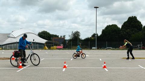 Ein älterer Radfahrer und ein jüngerer Radfahrer fahren auf Markierungen sowie um rote Verkehrshütchen herum. Eine Polizistin in Uniform gibt Anweisungen