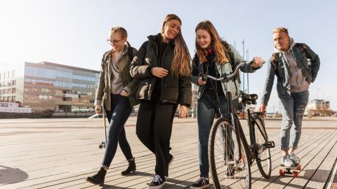 Vier Teenager sind auf einer Straße unterwegs. Ein Mädchen schiebt ein Fahrrad, ein Junge hat sich mit einem Skatebord an den Gepäckträger gehängt.