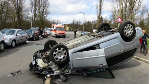 Ein schwerer Unfall auf einer Straße. Ein silbernes Auto liegt auf dem Dach, der Motorbereich ist zerstört. Im Hintergrund stehen weitere Fahrzeuge und ein Rettungswagen. Die Unfallaufnahme läuft.