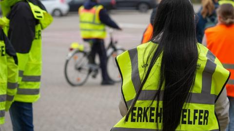 Eine Frau steht mit dem Rücken zur Kamera. Sie trägt eine gelbe Weste mit der Aufschrift "Verkehrshelfer". Im Hintergrund sind weitere Personen mit gelben und orangenen Westen zu sehen