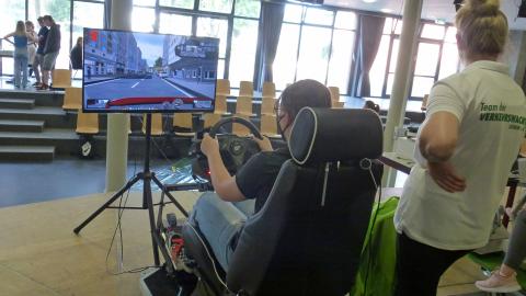 Eine junge Frau sitzt an einem Auto-Simulator. Eine Ausbilderin steht dahinter. Der Simulator ist offensichtlich in einer Aula aufgebaut