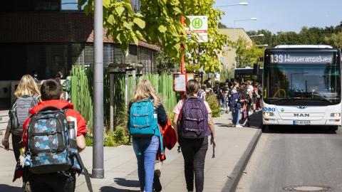 Mehrere ältere Schulkinder gehen auf einem Gehweg auf eine Bushaltestelle zu. Dort steht schon ein weißer Linienbus