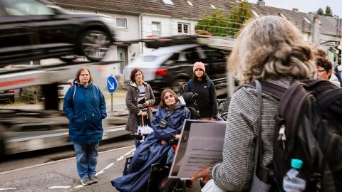 Ein Bild einer Szene im Straßenverkehr mit einer Frau im Rollstuhl. Es findet eine Beratung statt.