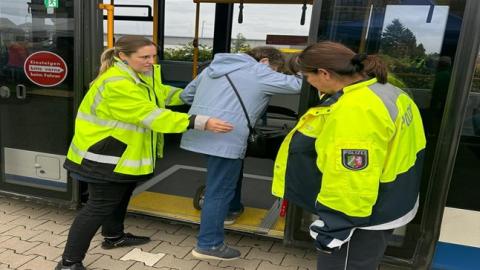 Bild einer Seniorin mit einem Rollator beim Einstieg in einen Linienbus. Rechts und links der Tür stehen zwei Polizistinnen