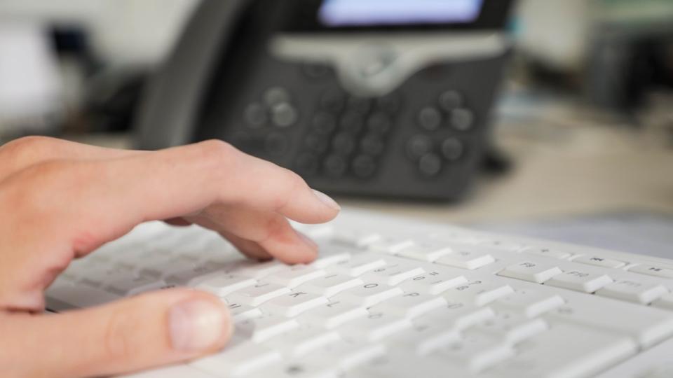 Eine Hand tippt auf einer Computertastatur. Im Hintergrund steht ein Telefon.