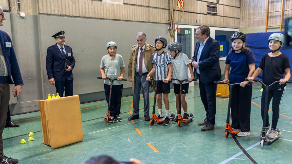 Die Minister Reul und Krischer besuchen einen Stand an dem mit Rollern die Verkehrssicherheit geübt wird. Das ganze in einer Turnhalle
