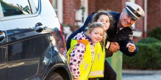 Ein Polizeibeamter steht mir zwei Schulkindern hinter einem Auto und zeigt auf die Straße