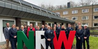 In der Mitte des Bildes stehen die drei Buchstaben NRW in den Landesfarben grün, weiß und rot. Die Mitglieder der Projektgruppe stehen neben und hinter dem Schriftzug, der Projektleiter davor