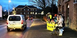 Auf einem Gehweg stehen in der Dämmerung Schülerinnen und Schüler sowie Eltern, alle tragen gelbe Warnwesten und Reflektoren. Sie halten ein Banner, den Schriftzug kann man nicht erkennen. Im linken Bildrand fährt ein Auto vorbei