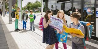 Mehrere Schulkinder stehen an einer Bushaltestelle. Sie haben Unterrichtsmaterial in der Hand. Im Hintergrund steht ein Linienbus