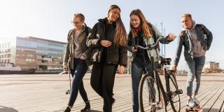 Vier Teenager sind auf einer Straße unterwegs. Ein Mädchen schiebt ein Fahrrad, ein Junge hat sich mit einem Skatebord an den Gepäckträger gehängt.