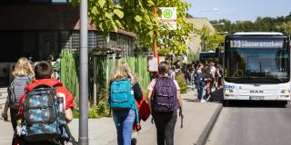 Mehrere ältere Schulkinder gehen auf einem Gehweg auf eine Bushaltestelle zu. Dort steht schon ein weißer Linienbus